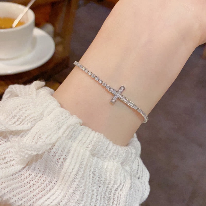 Silver-tone sparkling cross tennis bracelet worn on a wrist beside a cream sleeve.