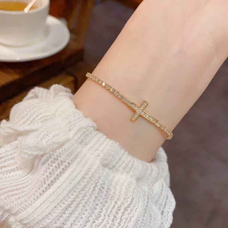 Gold-tone sparkling cross tennis bracelet worn on a wrist beside a cream sleeve.